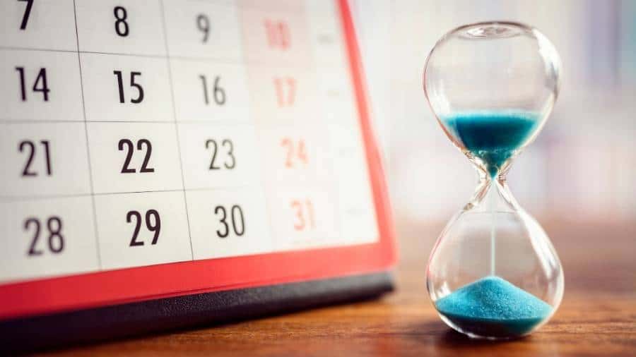 Clear hourglass with blue sand in front of a propped-up calendar. 