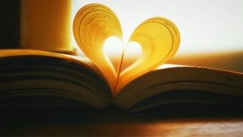 An open book with the left and right pages folded in toward the book's center to form a heart shape. 