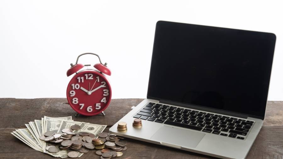 A laptop, a red alarm clock, and a pile of bills and coins sit on a dark-brown wooden bench. 