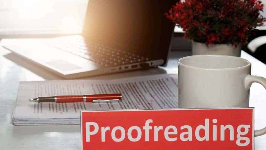 A laptop, coffee mug, and a pile of papers that have been proofread with a red pen sit on top of a desk in a home office. 