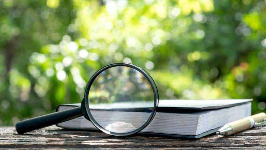 A magnifying glass, book, and writing pen lying on a wooden bench outside. 